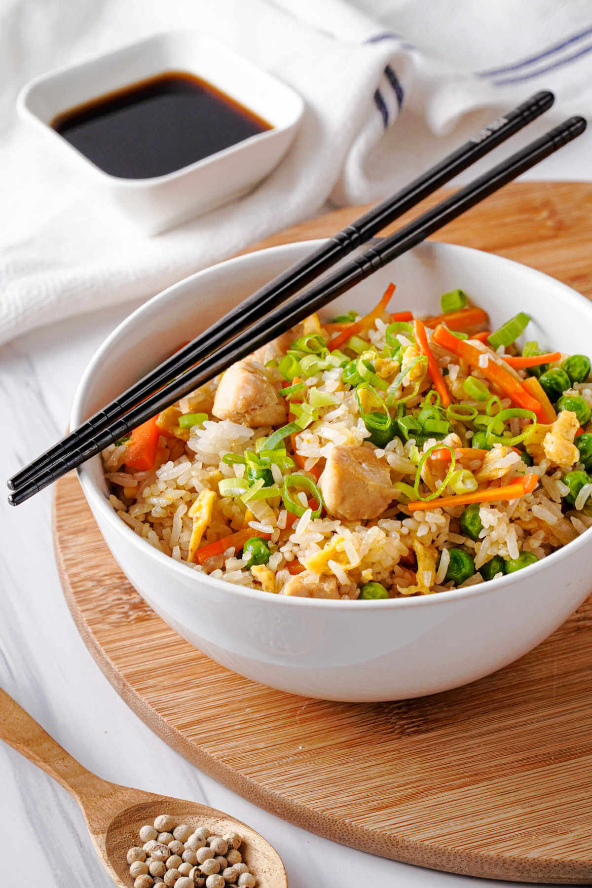 chicken fried rice in a white bowl with chopsticks and a bowl of soy sauce in the background sitting on a wooden board.