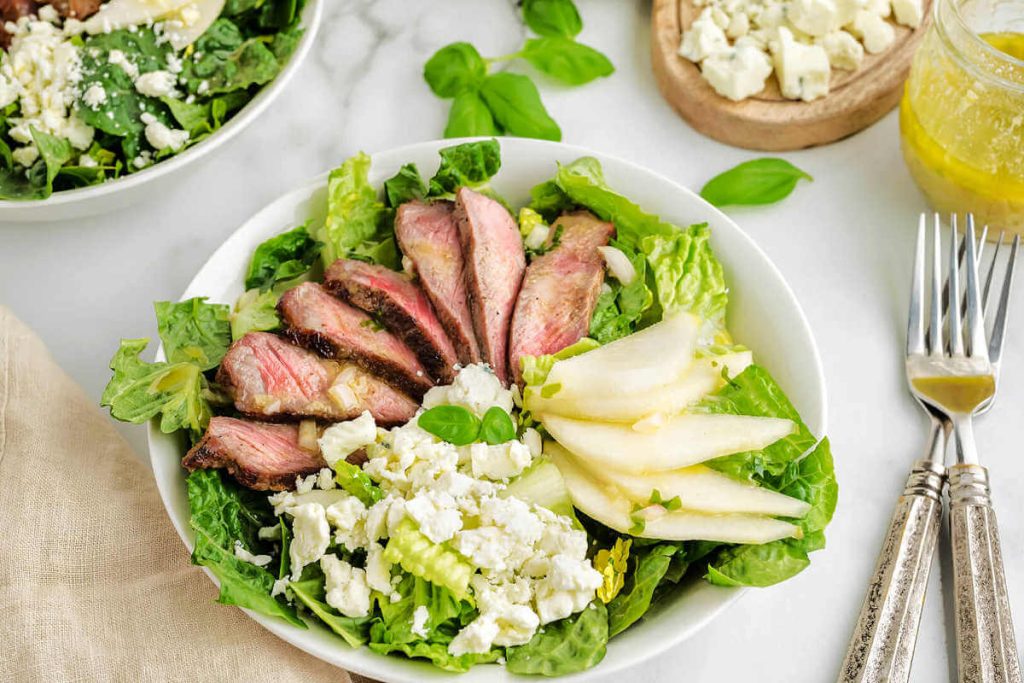 Grilled Steak Salad with a French Vinaigrette in a white bowl on a table with condiments and silverware.