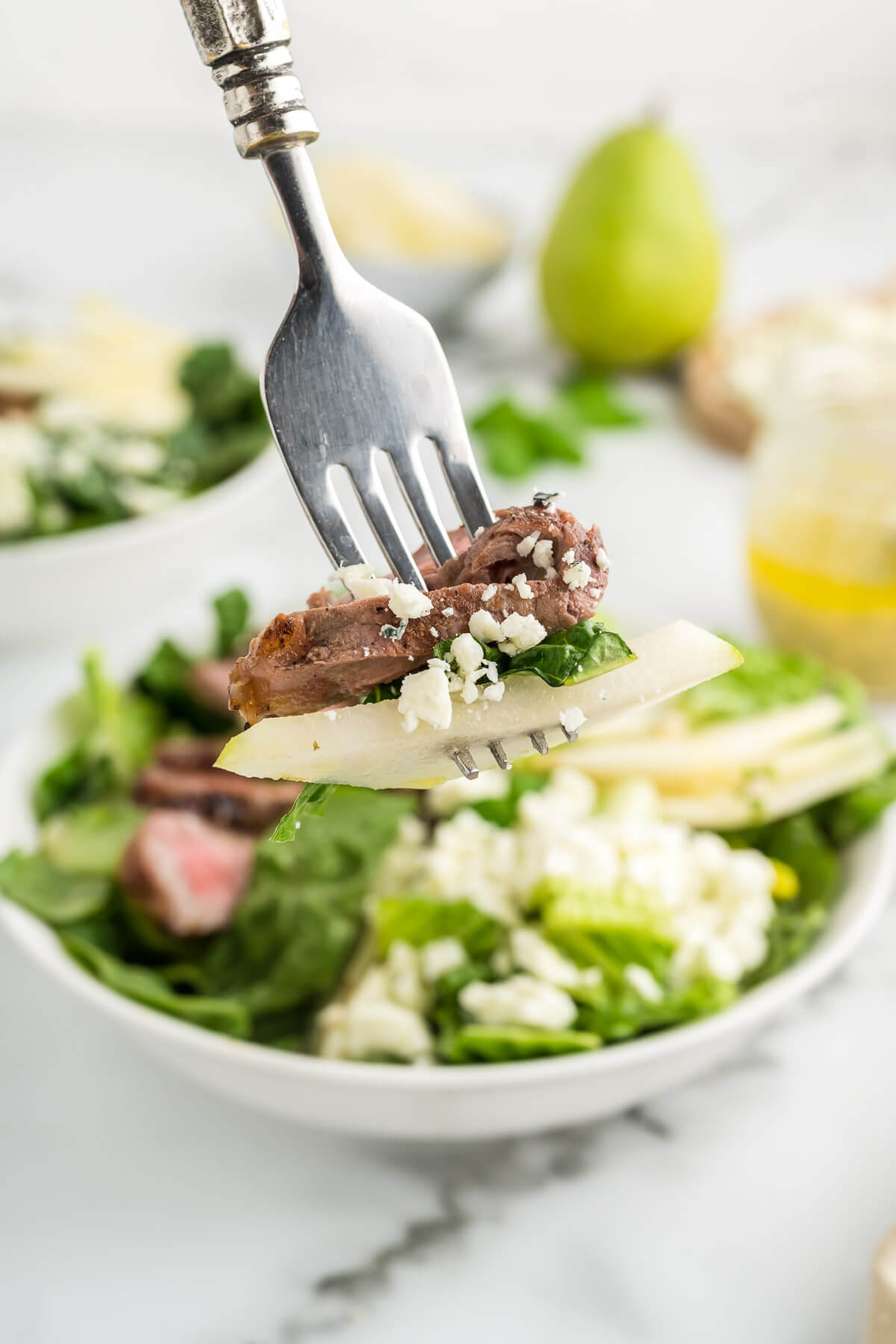 a bite of steak and pear on a fork with a bowl of salad in the background.