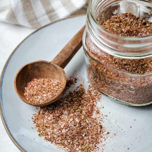 Homemade steak seasoning in a jar sitting on aplate with a wooden spoon.