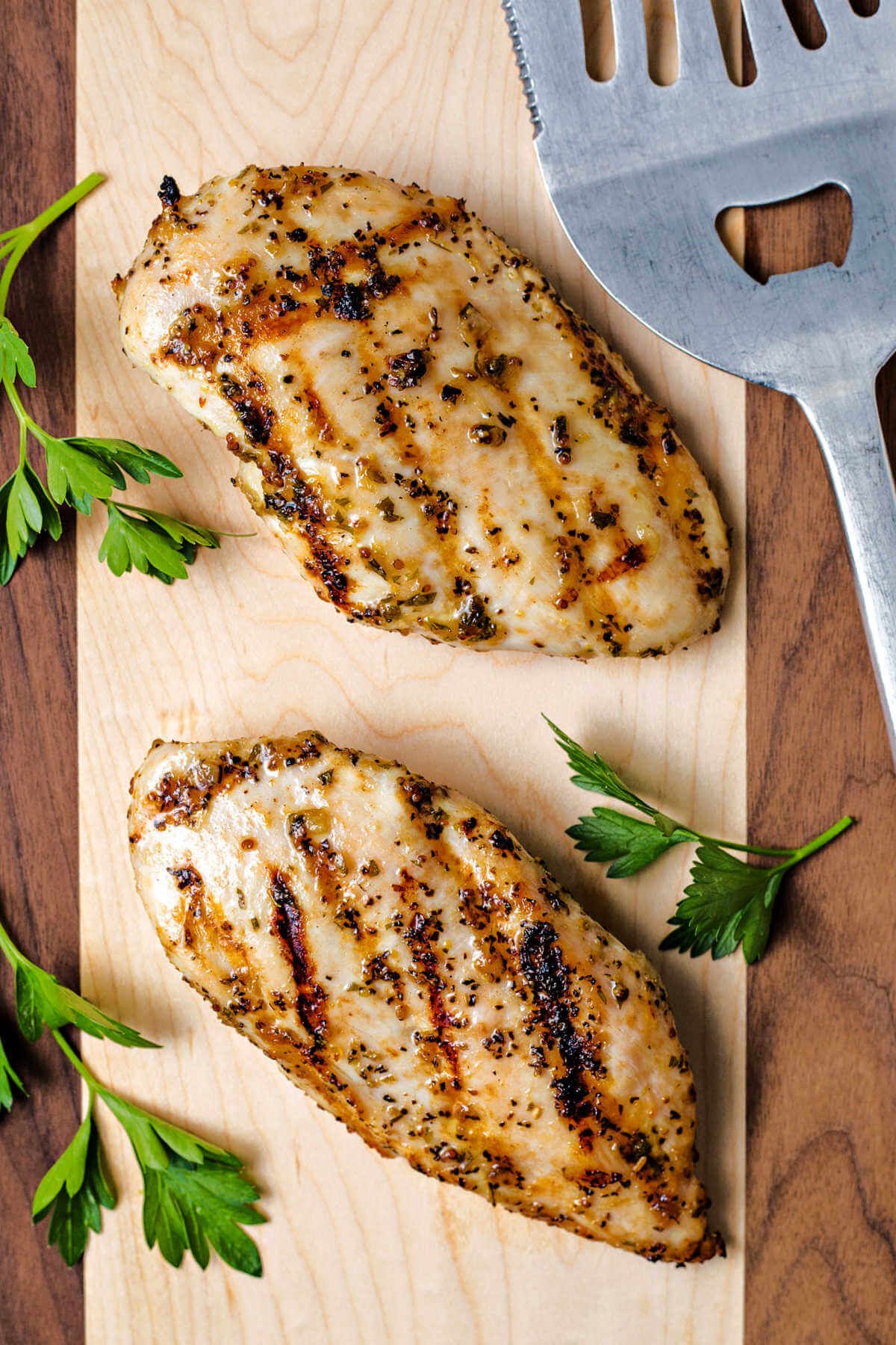 two Italian grilled chicken breasts on a wooden cutting board with a spatula to the side.