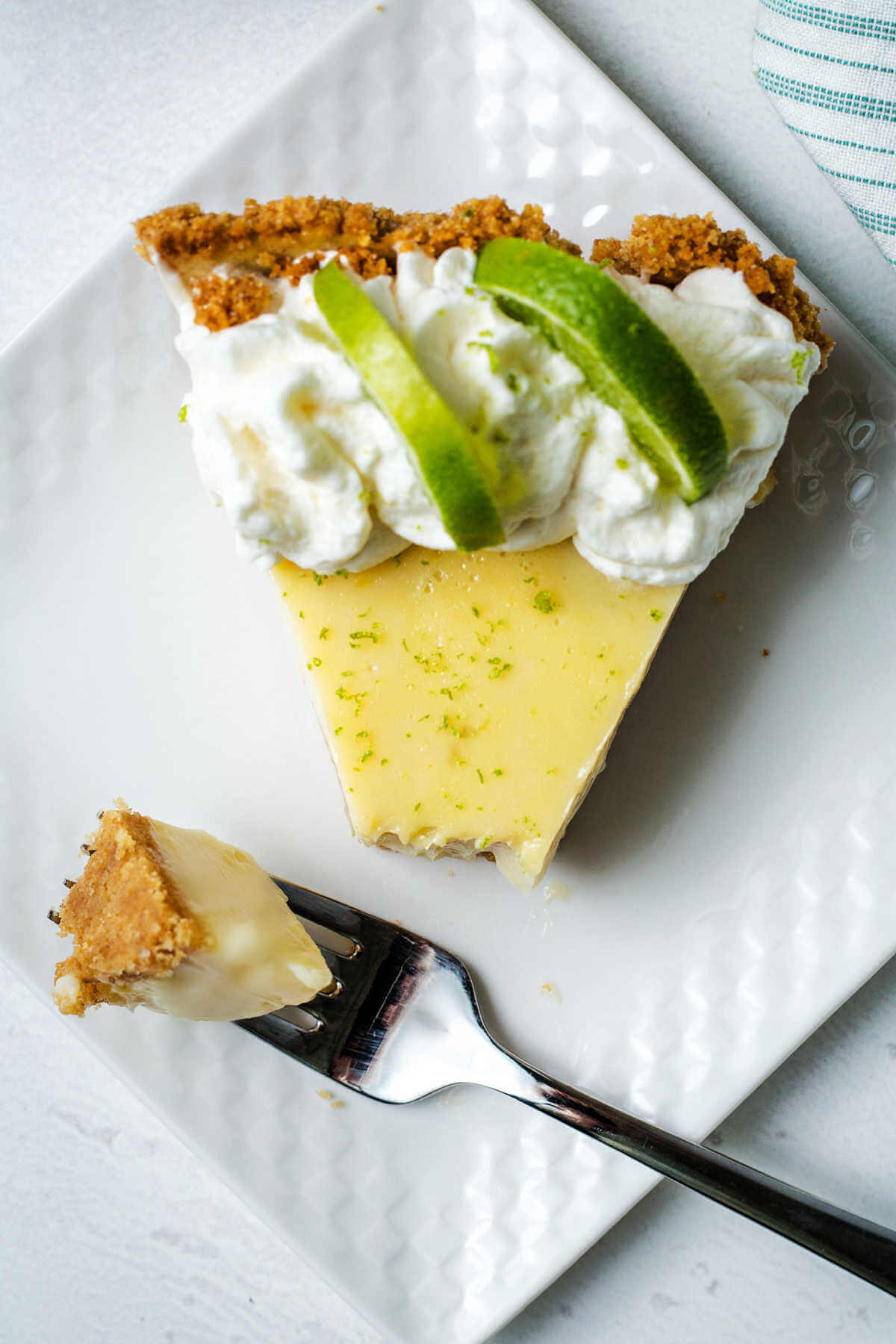 a bite of key lime pie on a fork on a plate with a piece of partially eaten pie.