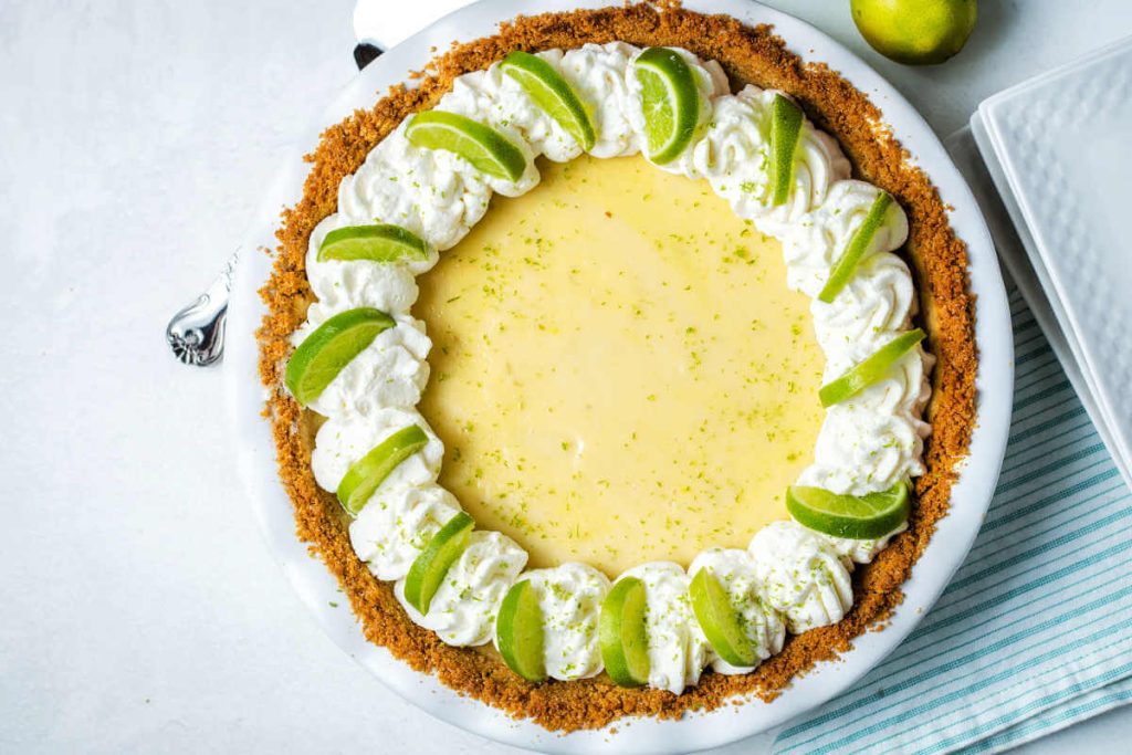 a whole key lime pie garnished with fresh whipped cream and lime slices on a table with limes.