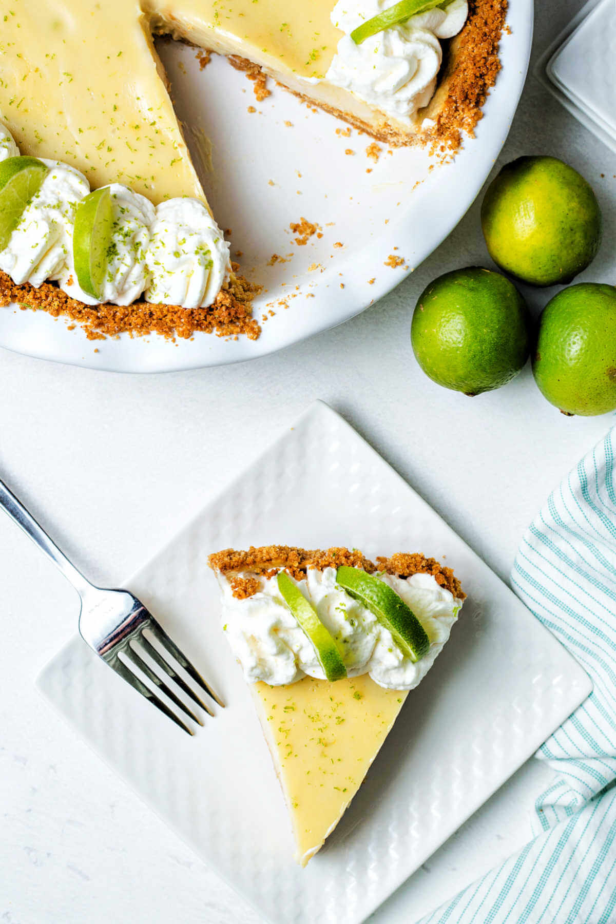 a slice of key lime pie on a plate with the remaining pie in the background with three limes on a table.