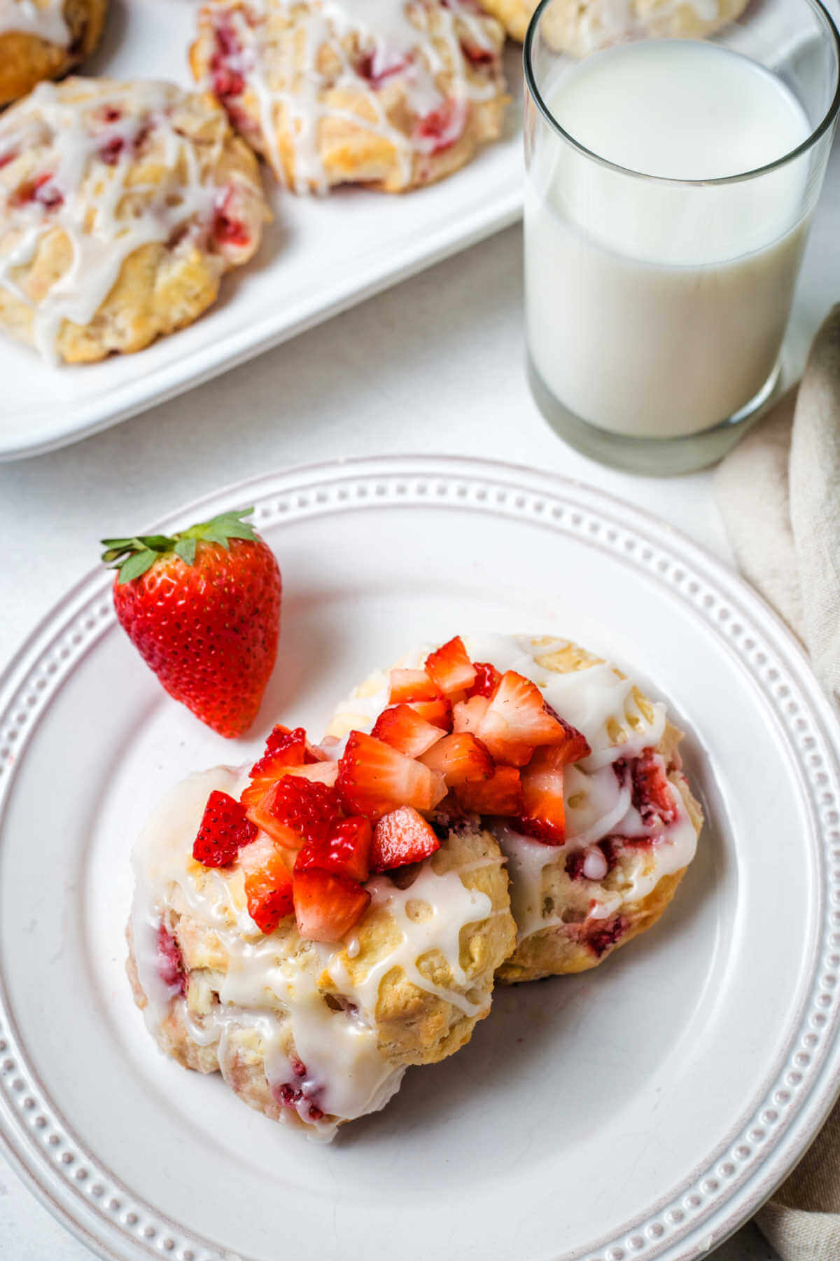 two strawberry biscuits on a white plate with more diced strawberries ladled on top and a glass of milk in the background.