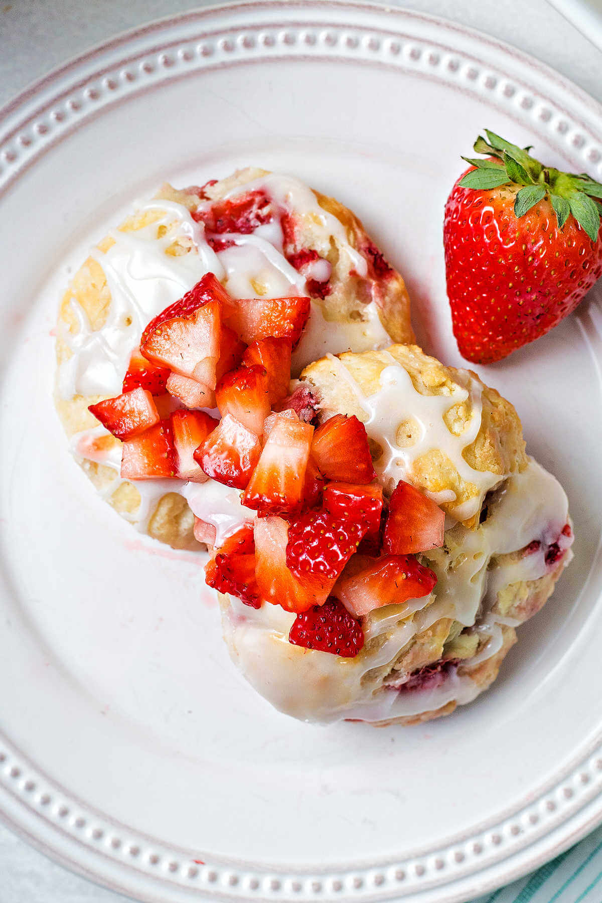 two strawberry biscuits on a white plate with more diced strawberries ladled on top and a whole strawberry to the side.