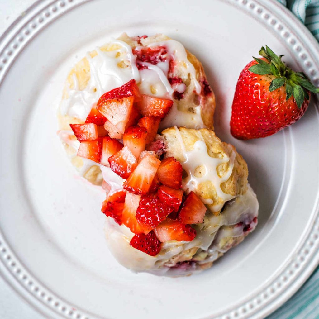 two strawberry biscuits on a white plate with more diced strawberries ladled on top and a whole strawberry to the side.