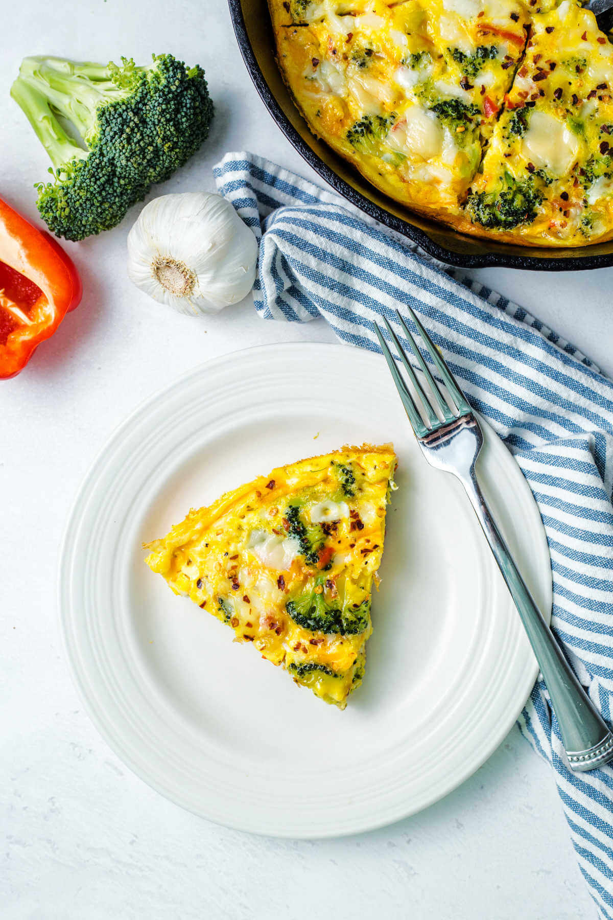 a serving of healthy vegetable frittata on a white plate with a cast iron skillet with more frittata in the background on a table.
