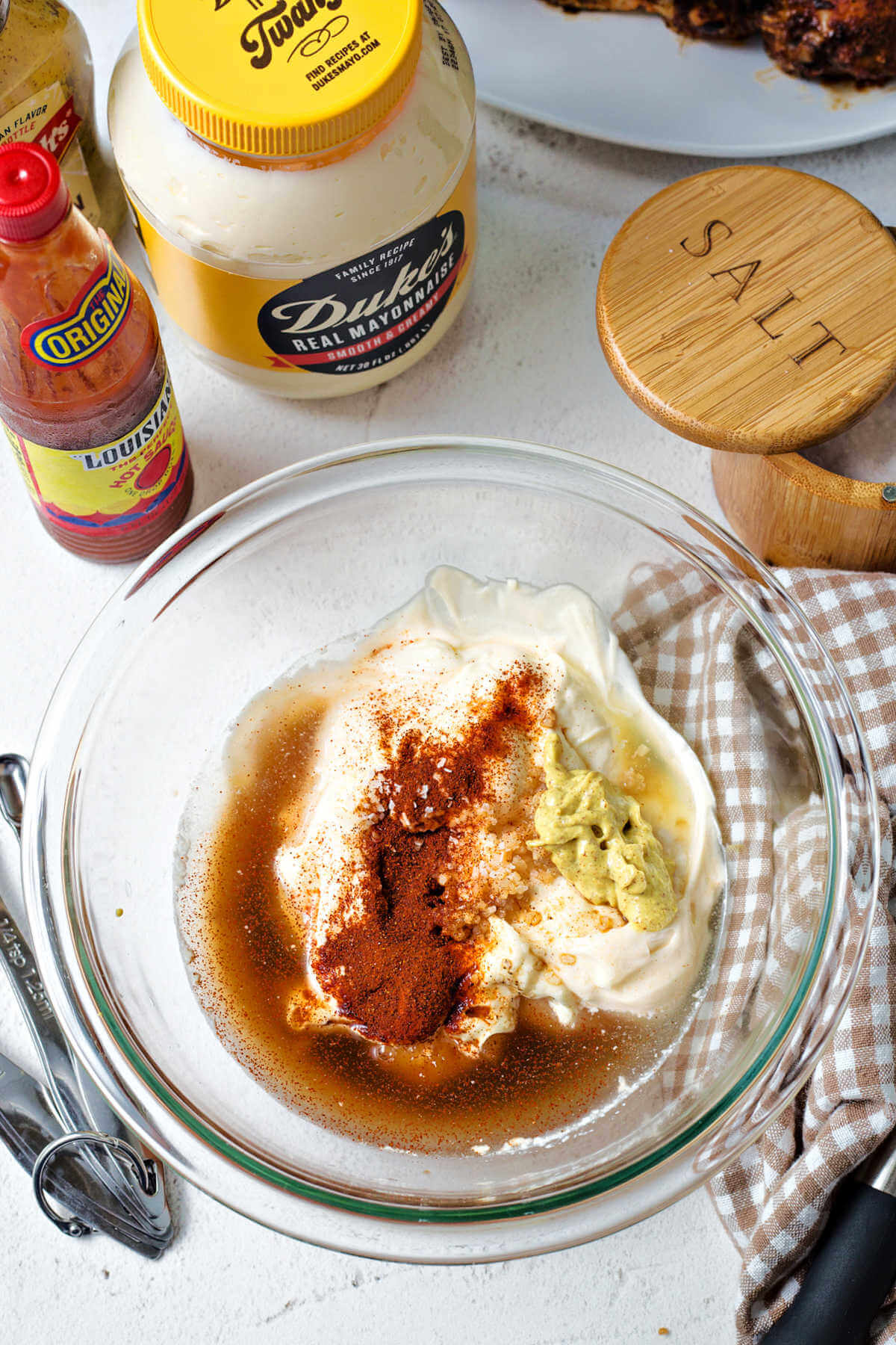 ingredients for Alabama White Sauce in a glass bowl on a table with jars of mayo and hot sauce in the background,