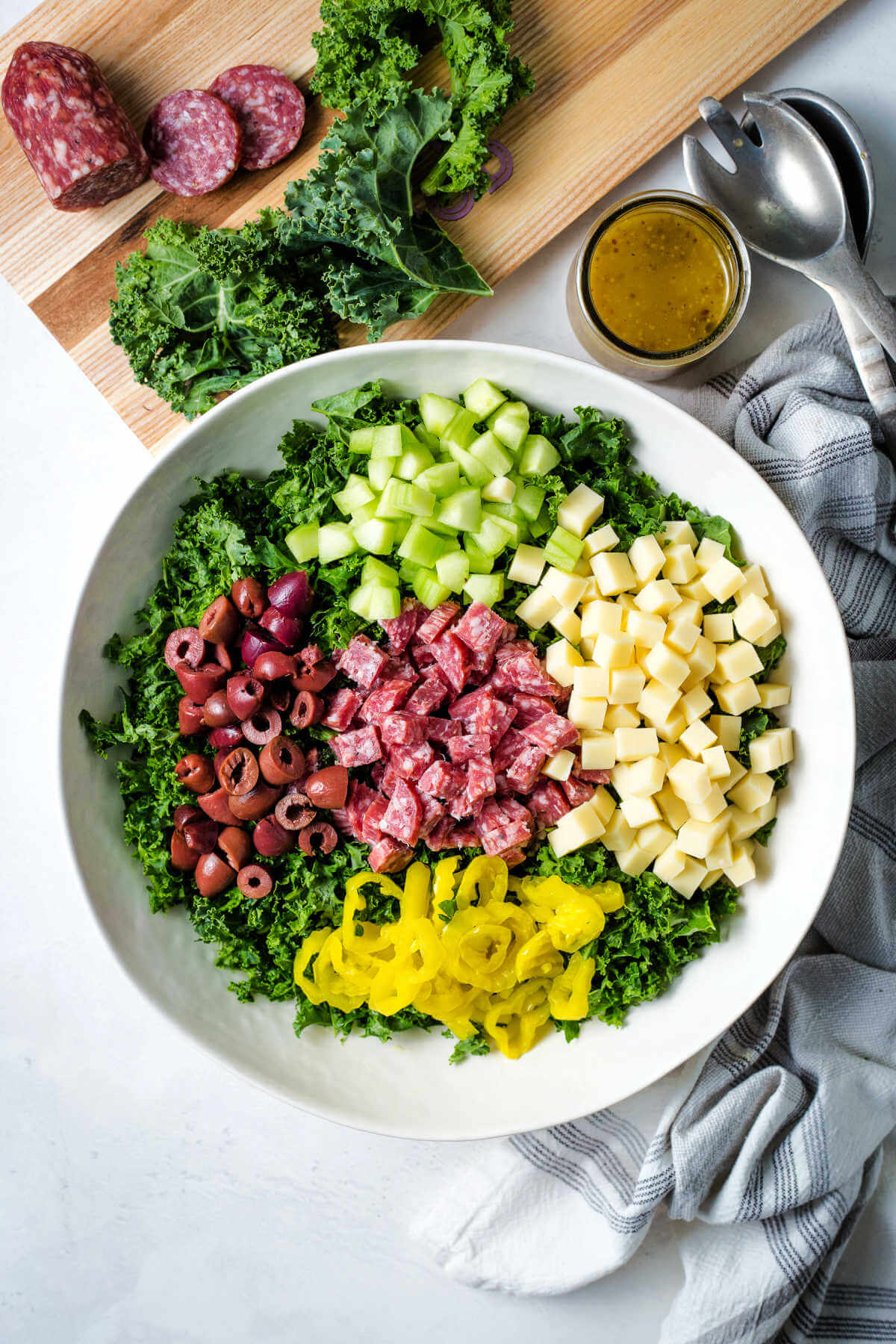 chopped ingredients in a bowl and vinaigrette in a bottle for chopped kale salad on a table with a cutting board with salami and kale leaves.