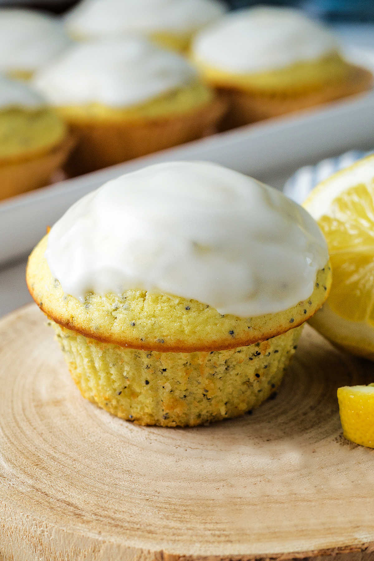 a glazed lemon poppyseed muffin on a wooden block wit a tray of muffins in the background.