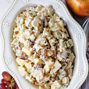Southern chicken salad with grapes and pecans in a decorative white bowl on a table with a cluster of grapes and an apple.