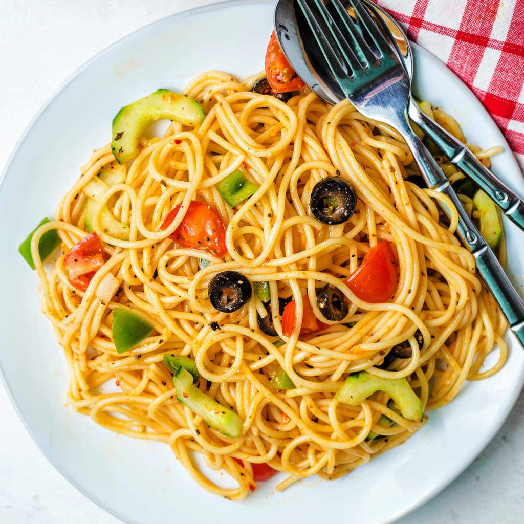 a serving of spaghetti pasta salad on a plate with a fork and spoon and a red checked napkin on a table.