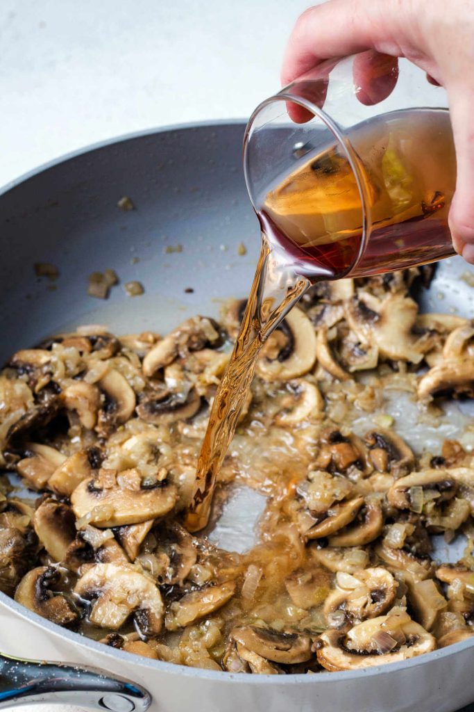 pouring wine into a sizzling pan of cooked mushrooms.