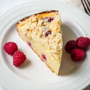a slice of raspberry ricotta cake on a plate with raspberries on a table with a cake stand in the background.