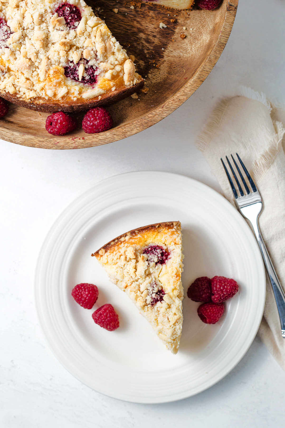 top down view of a slice of raspberry ricotta cake on a plate with raspberries on a table with a cake stand in the background.