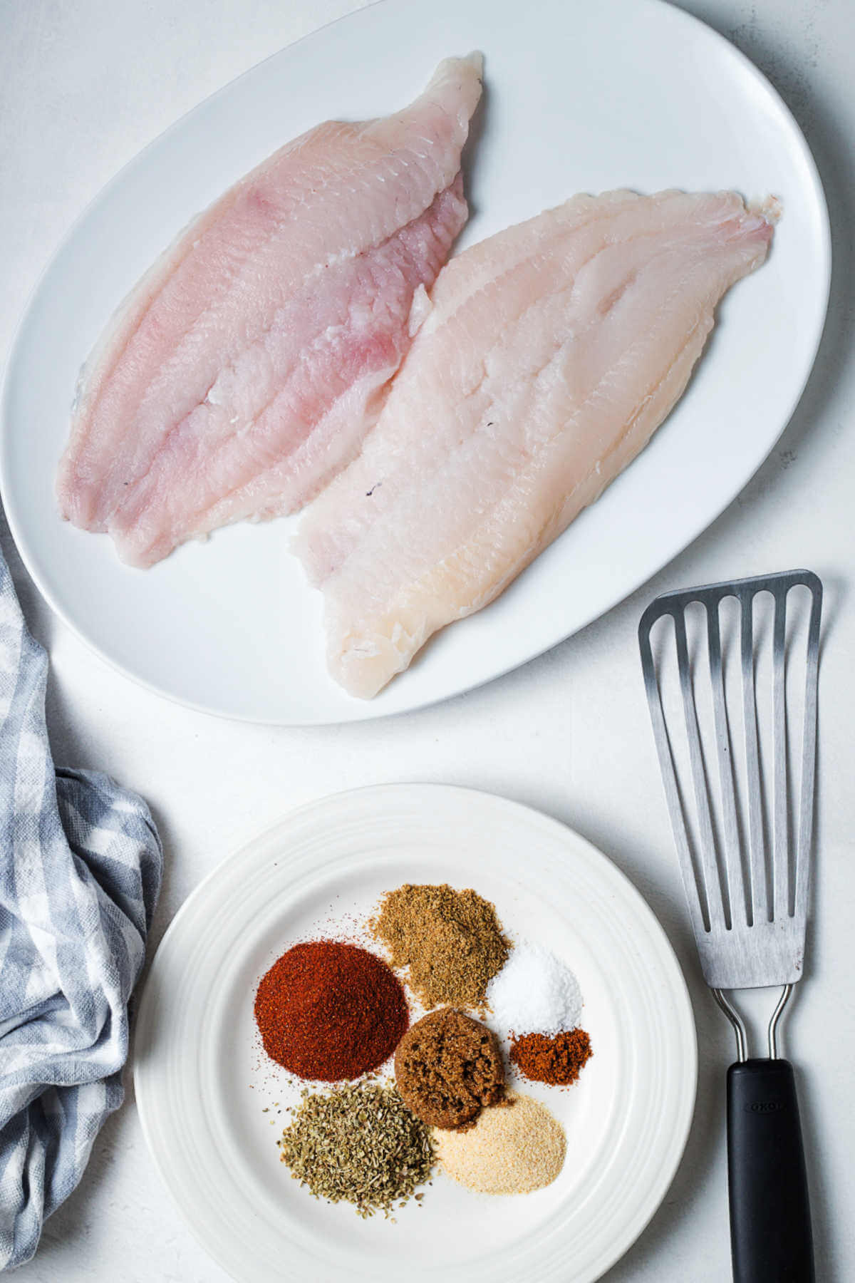 catfish filets on a plate with a fish turner and a plate of spices for making blackened fish.