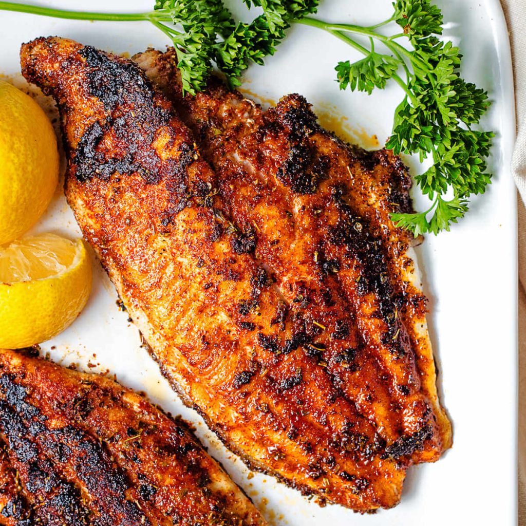 a blackened catfish filet on a plate with parsley and lemon wedges.