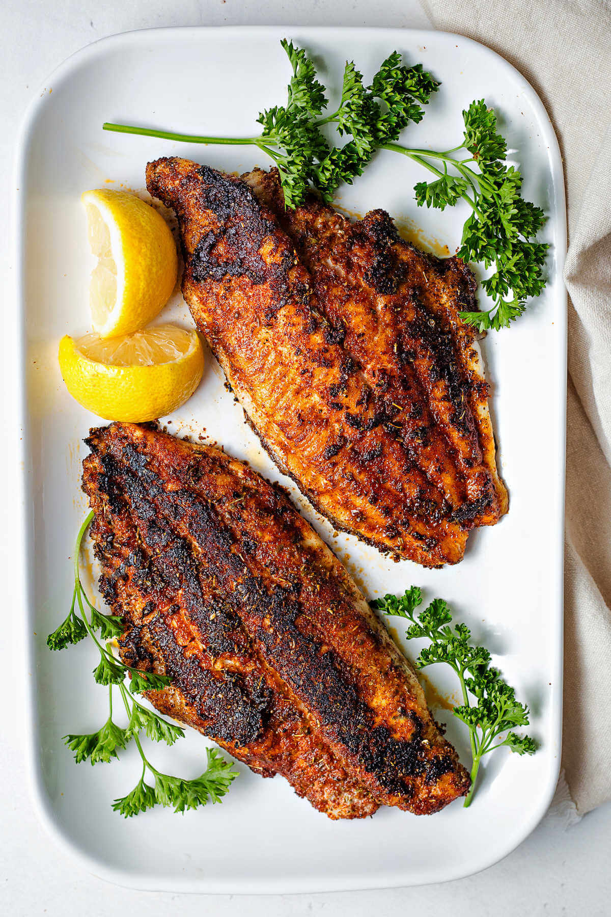 two blackened catfish filets on a white platter with lemon wedges and parsley.