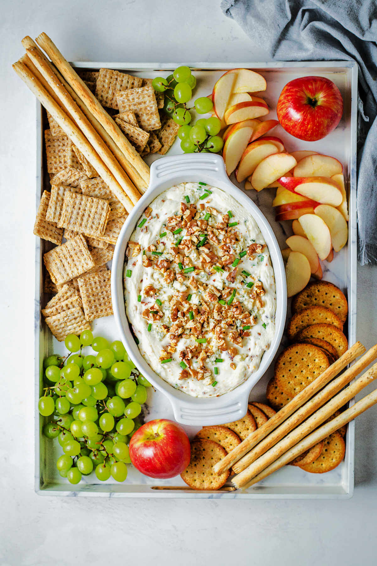 blue cheese dip with bacon, chives and walnuts on a tray arranged with bread sticks, assorted crackers, apple slices and grape clusters on a table.