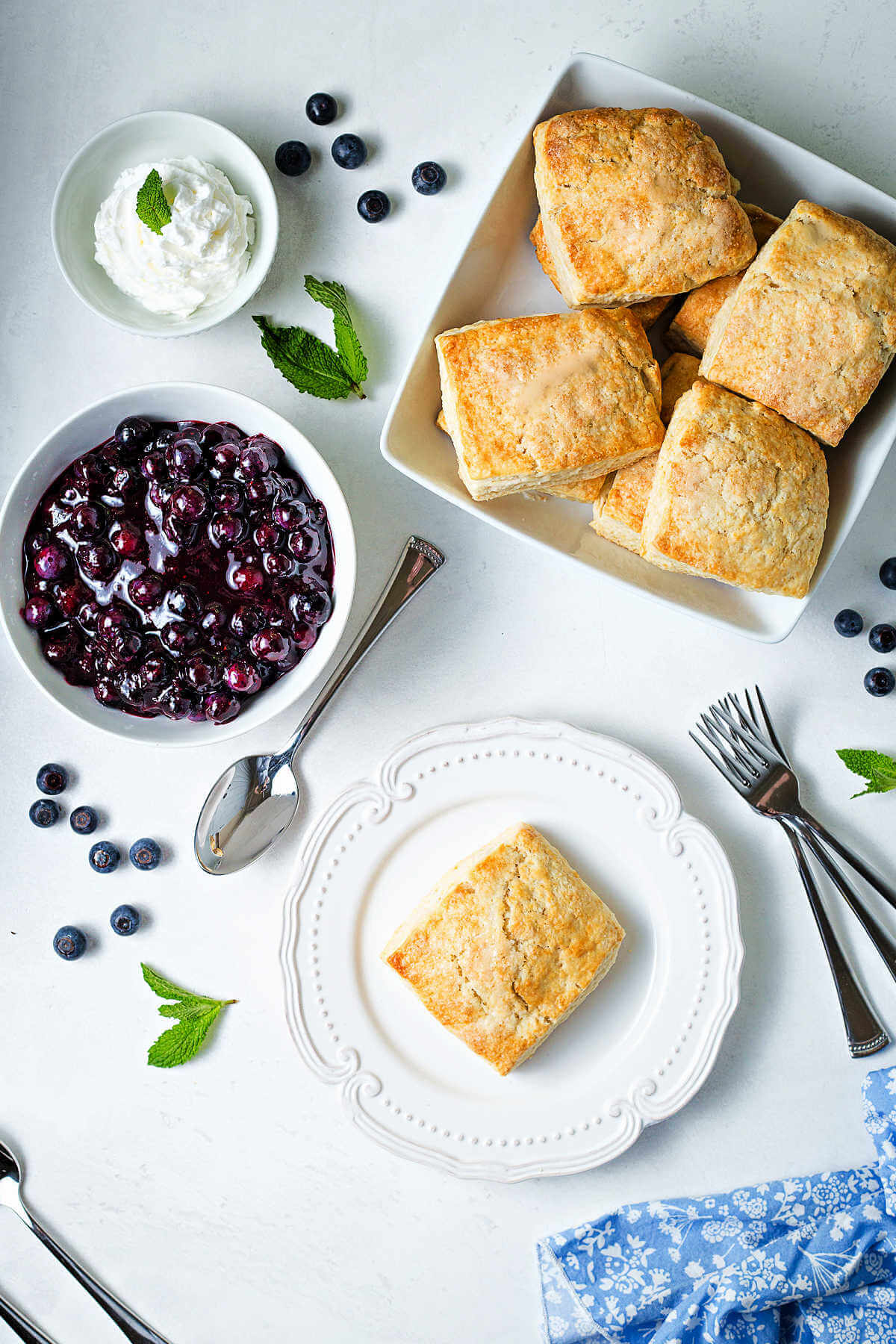baked shortcakes, a bowl of blueberries, and a bowl of whipped cream on a table.