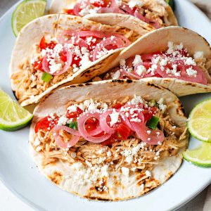 chicken street tacos on a plate garnished with salsa, pickled red onions, feta cheese and lime slices.