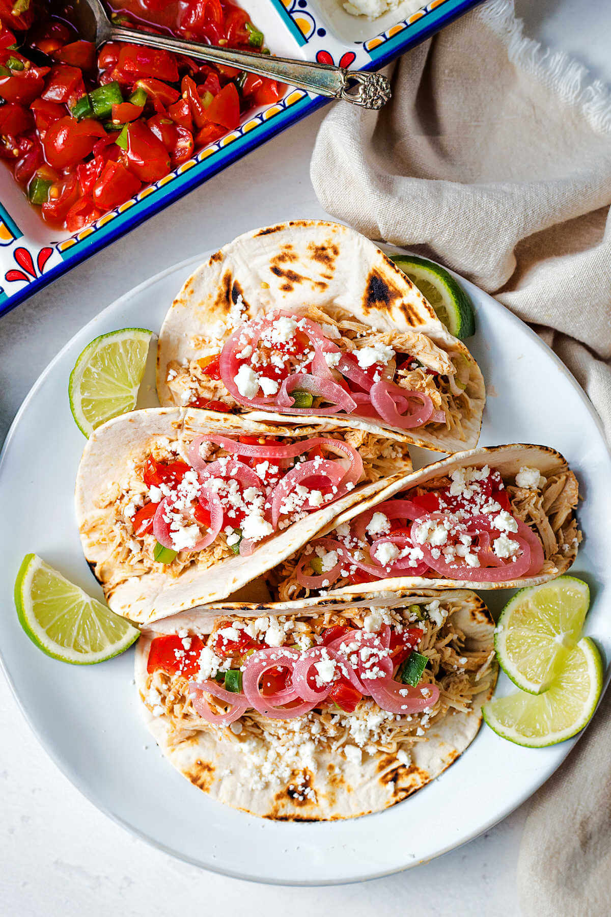 chicken street tacos on a plate garnished with salsa, pickled red onions, feta cheese and lime slices.