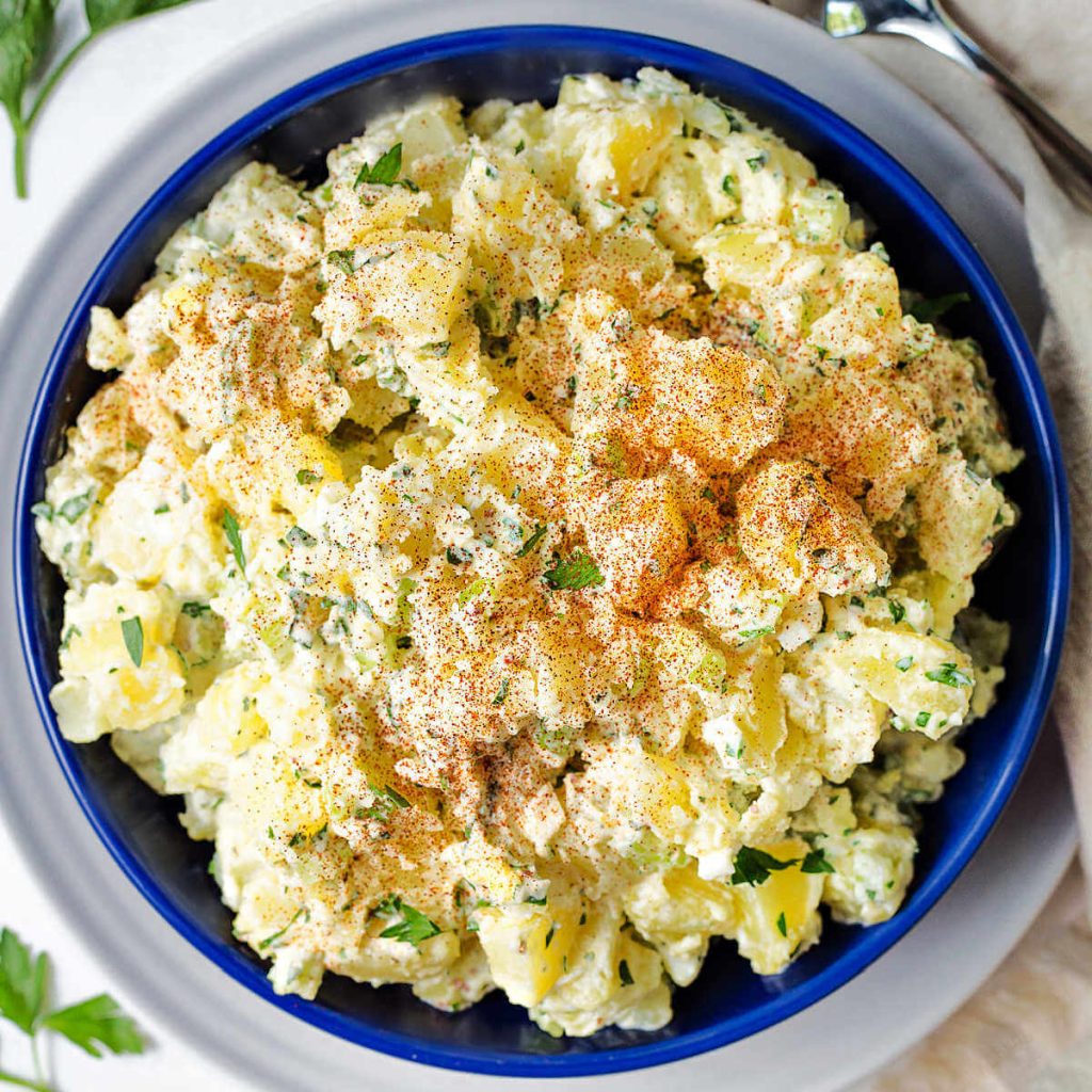 a bowl of fresh herb potato salad with paprika sprinkled on top on a table with a linen napkin and parsley sprinkled around.