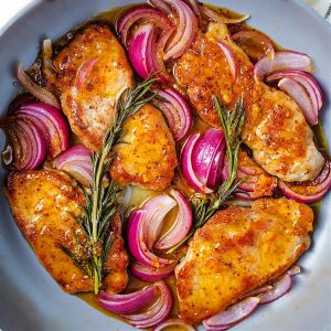 glazed pork chops in a skillet with red onions and rosemary sprigs.