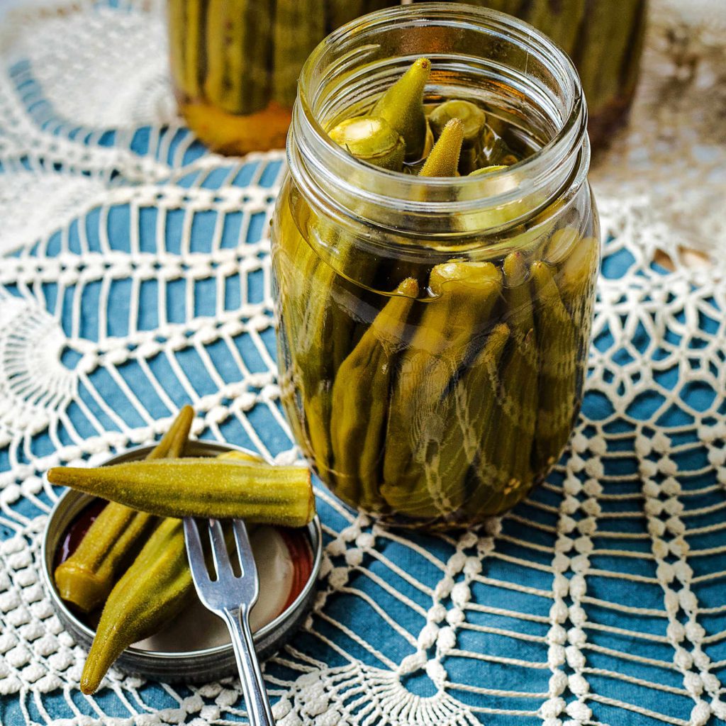 an open jar of pickled okra sitting on a table with a fork stuck into a pod of okra.