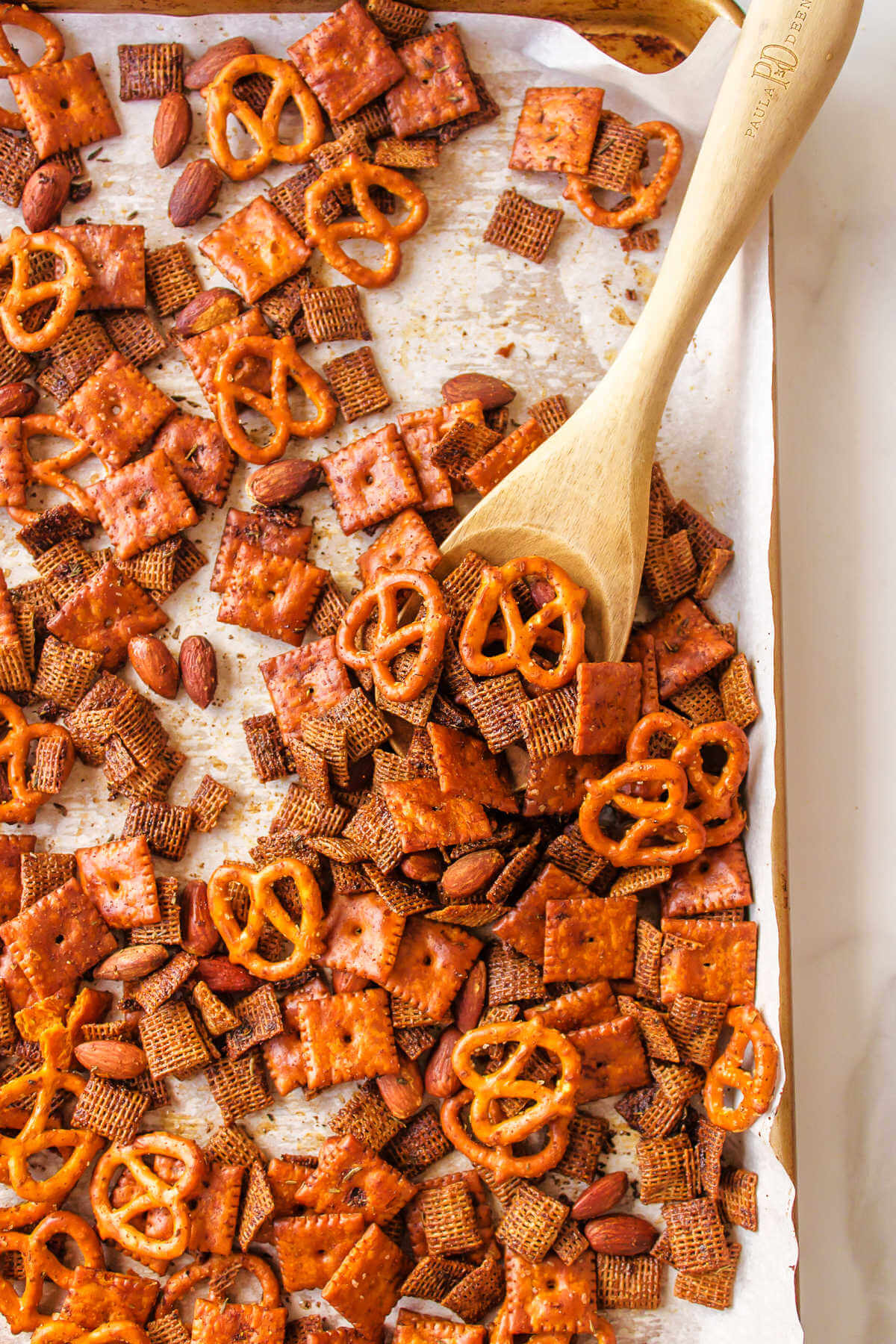 stirring Chex Mix on baking sheet with a wooden spoon.