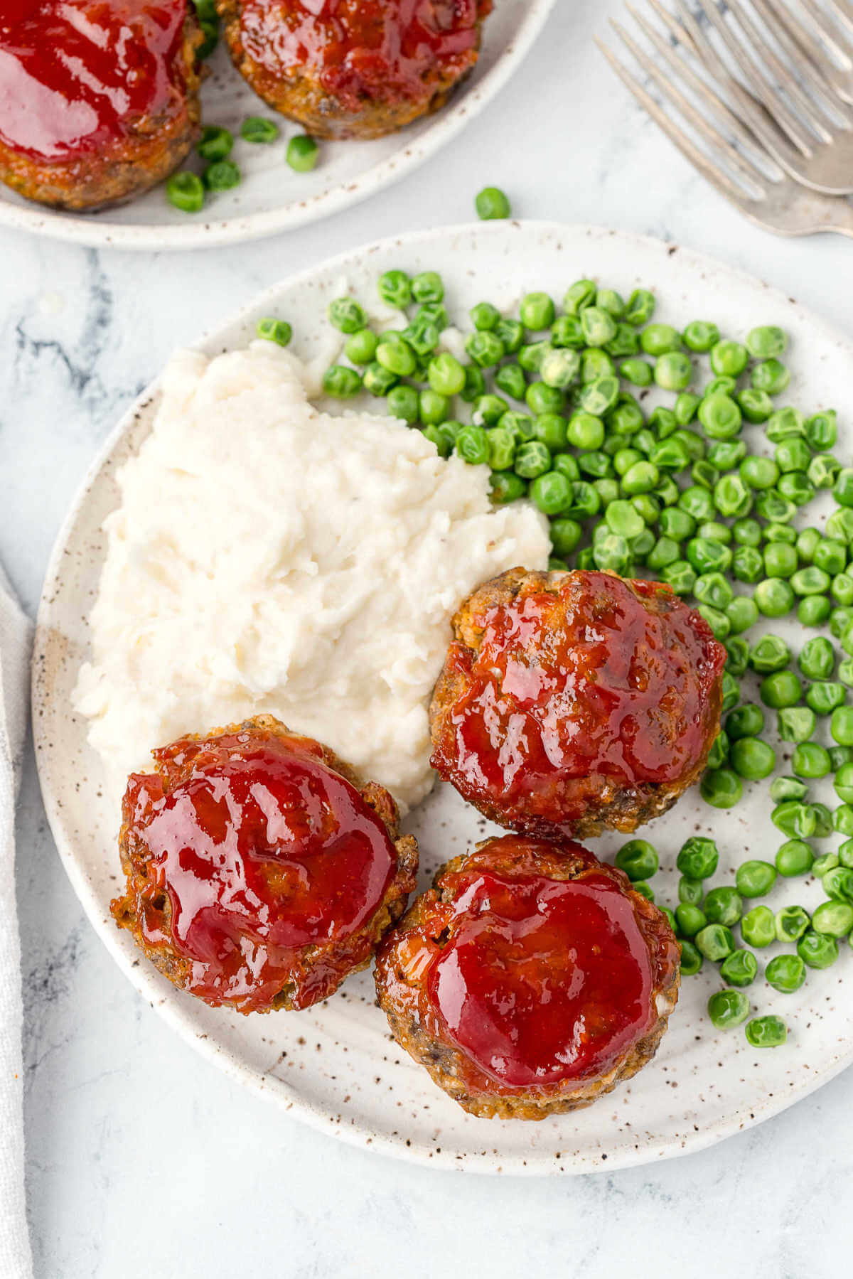 three meatloaf muffins on a plate with mashed potatoes and peas on a table.