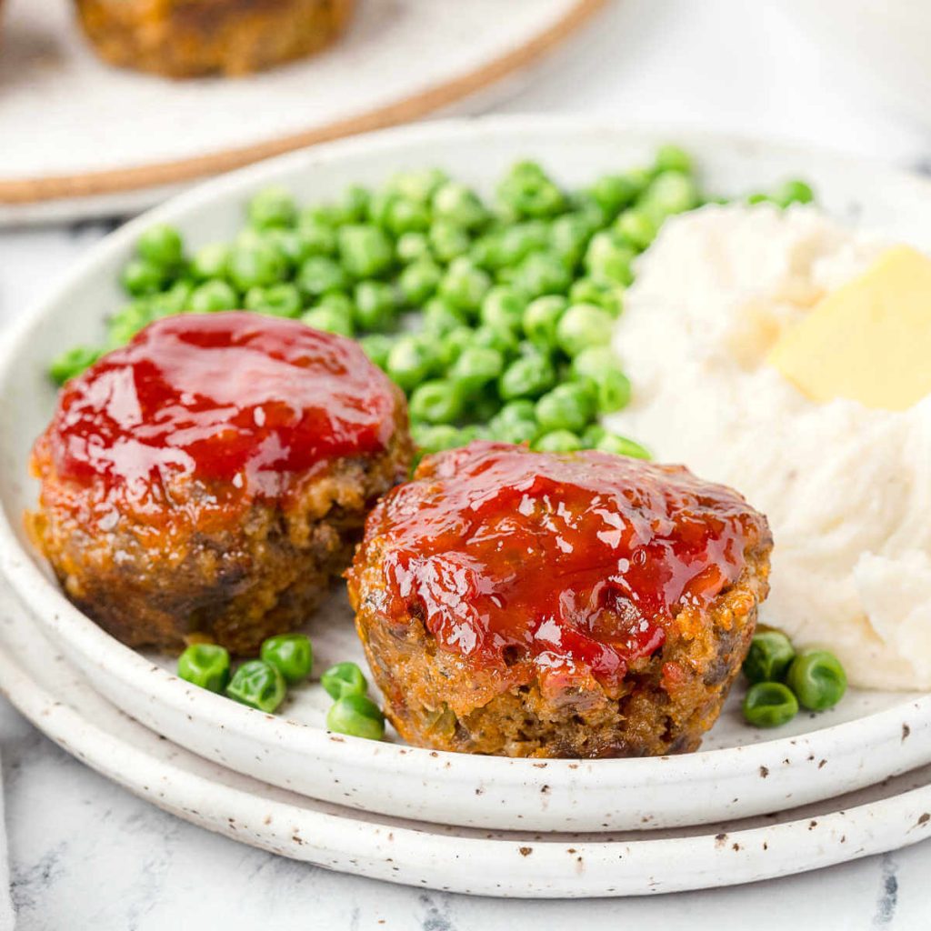 two mini meatloaf muffins on a dinner plate with peas and potatoes.
