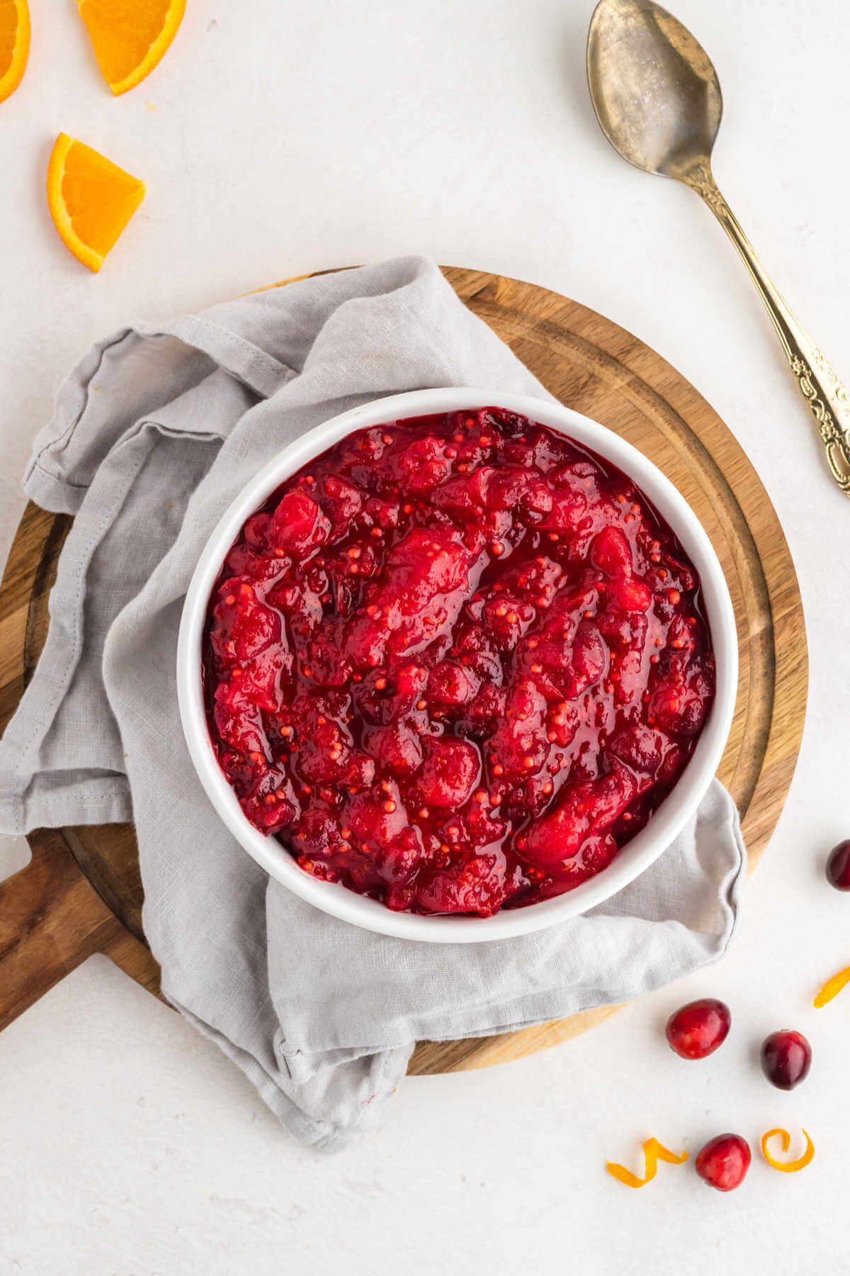 a bowl of cranberry chutney on a wooden trivet on a table scattered with cranberries and orange slices.