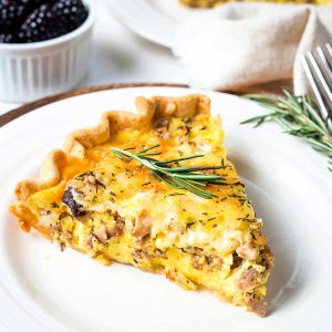 a slice of sausage and mushroom quiche with a rosemary sprig garnish on a plate on a table.