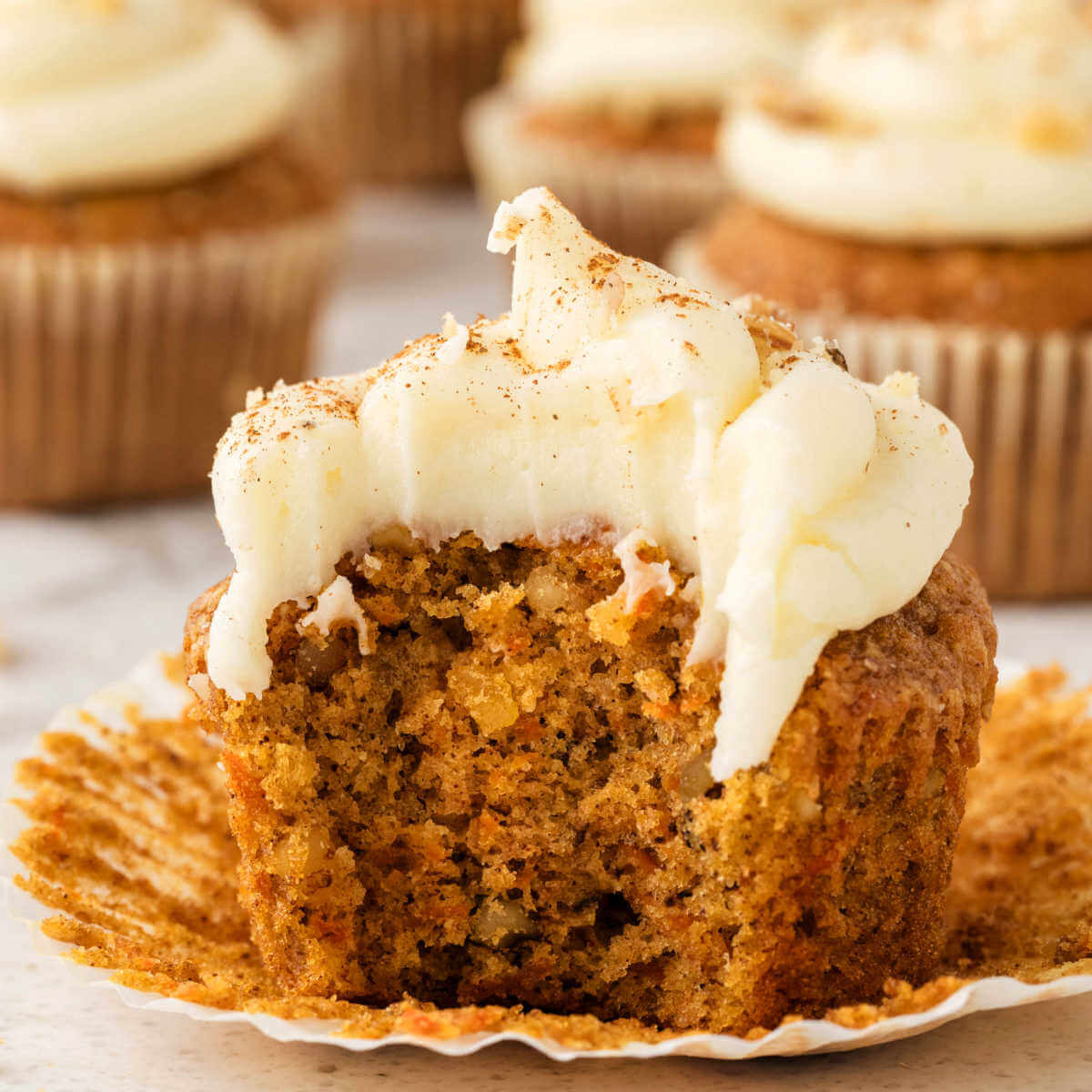 A carrot muffin with cream cheese frosting with a bite missing on a paper liner.