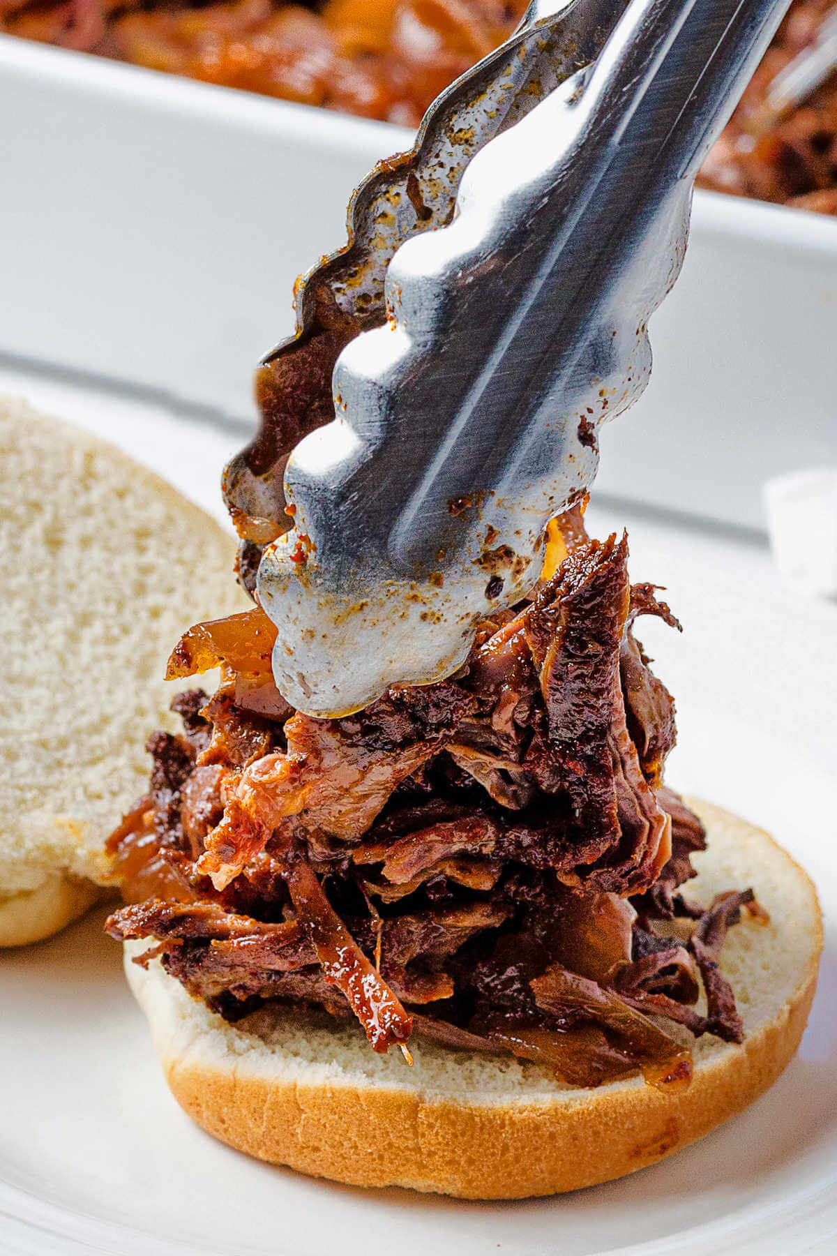 placing shredded beef on a bun with tongs on a plate.