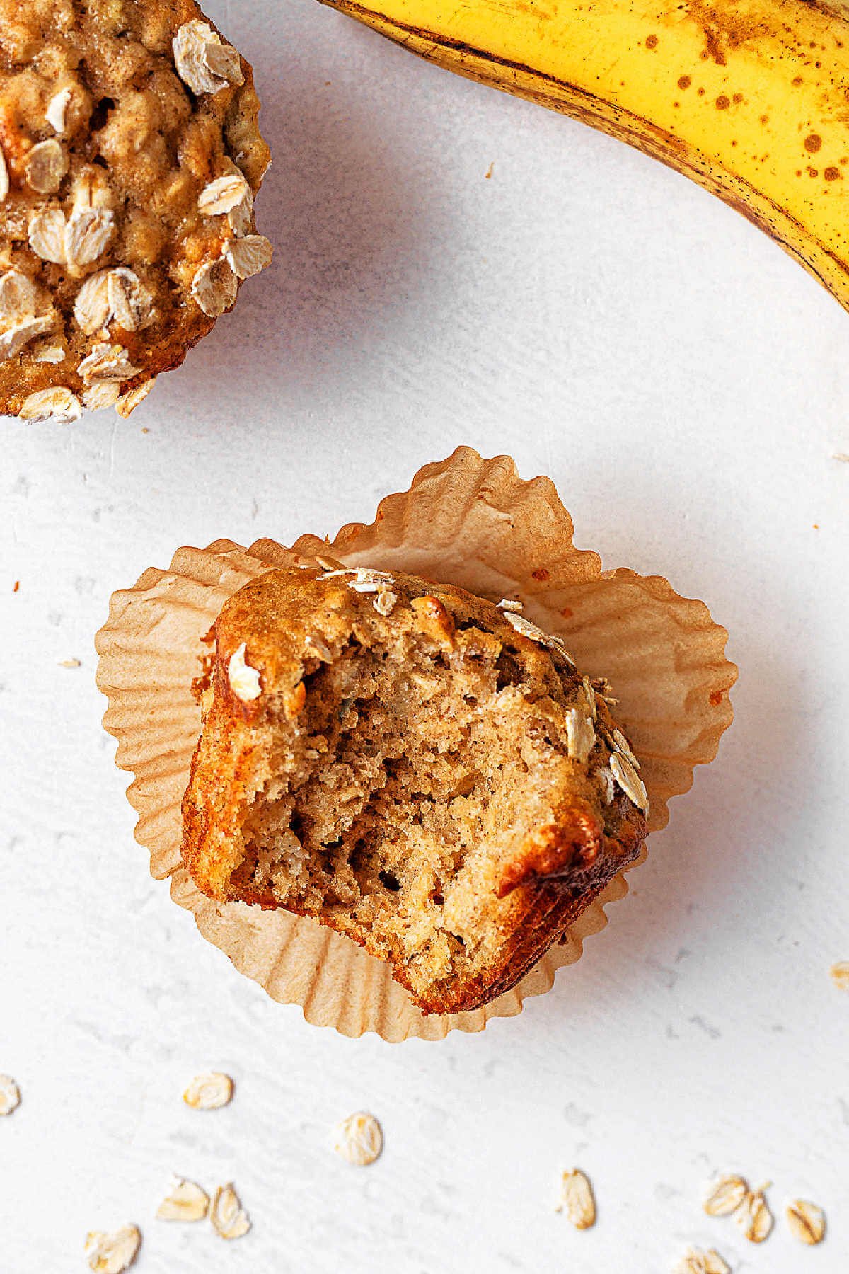 An Oatmeal Banana Muffin with a bite missing on top of a paper cupcake liner on a table.