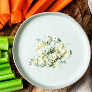 Blue cheese dressing in a bowl with blue cheese crumbles on top on a platter of carrot and celery sticks.
