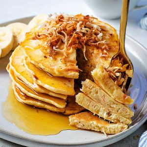 A stack of pancakes on a plate topped with toasted coconut with a fork spearing a bite.
