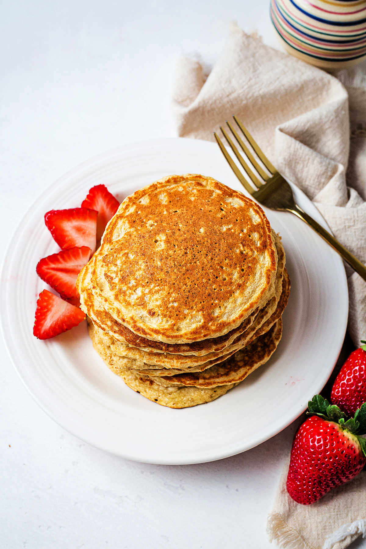 A stack of oat flour pancakes on a plate with sliced strawberries and a fork on the side.