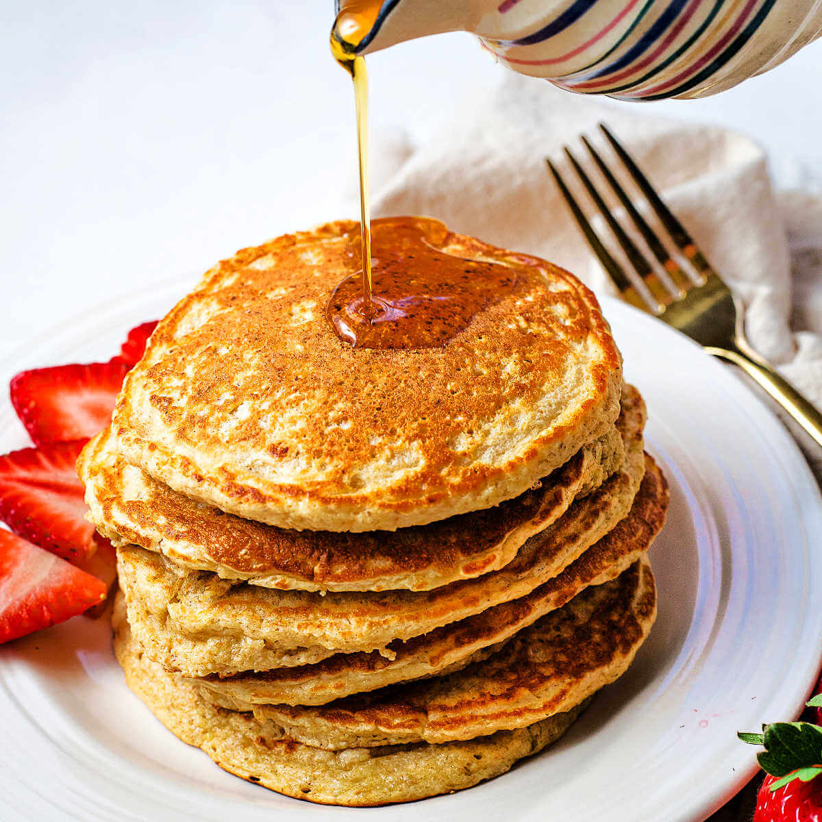 Pouring syrup over a stack of oat flour pancakes on a plate.
