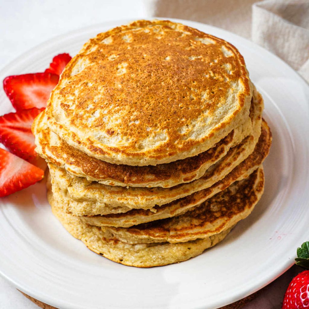 A stack of oat flour pancakes on a plate with sliced strawberries and a fork on the side.