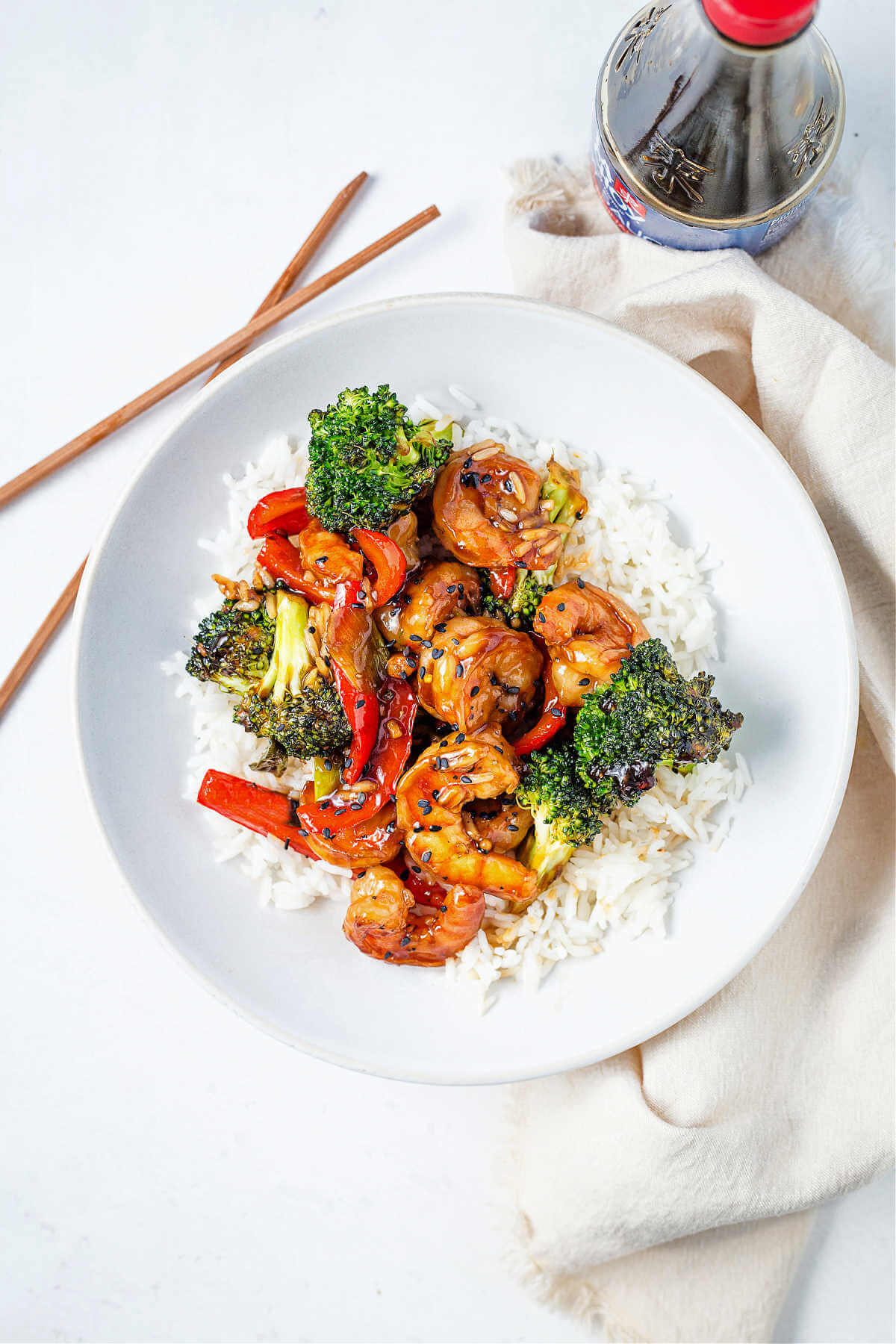 shrimp stir fry over a bed of white rice in a bowl on a table with chopsticks and a bottle of soy sauce.