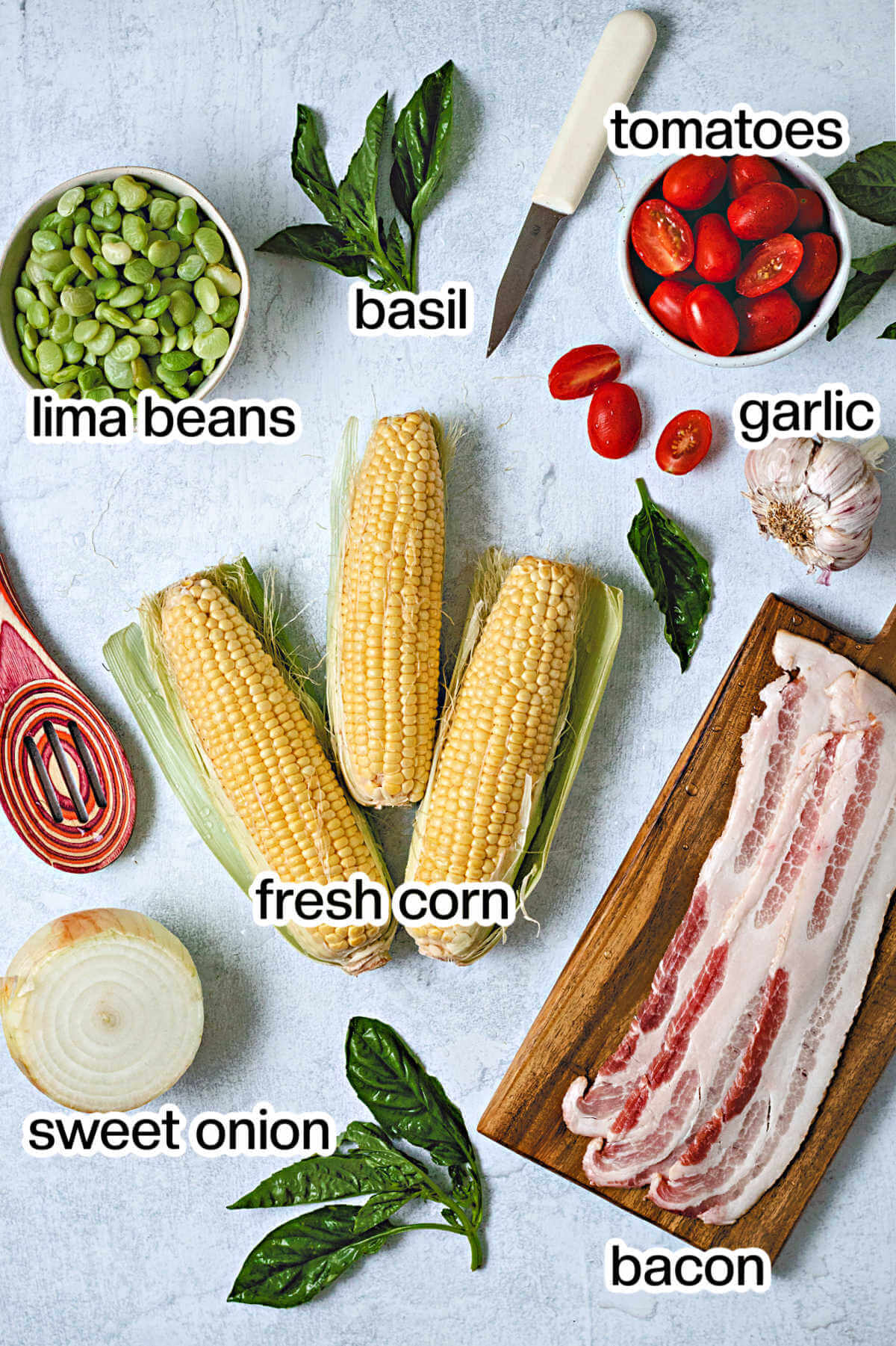 Ingredients for corn succotash on a table: 3 ears of corn, fresh basil, a bowl of lima beans, grape tomatoes, onion, garlic, and bacon slices.