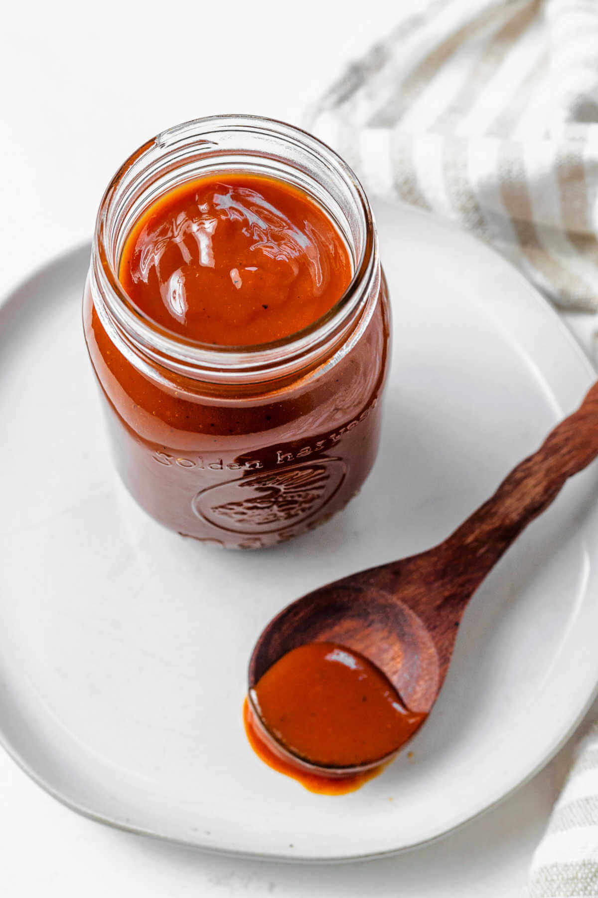 Barbecue sauce in a mason jar on a plate with a spoon of BBQ sauce resting on a plate.