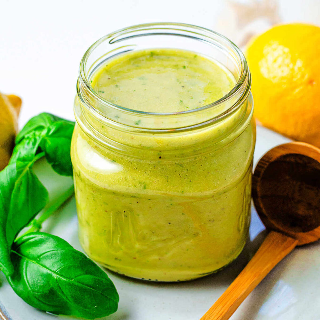 A jar of lemon basil vinaigrette on a table with fresh basil leaves, a lemon, and a wooden spoon to the side.