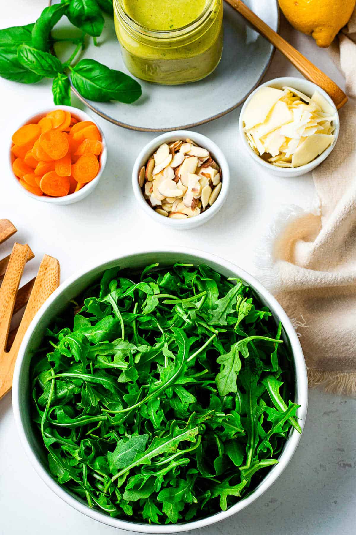 A bowl of arugula, sliced carrots, almonds, and shaved parmesan cheese with a jar of lemon basil dressing on a table.