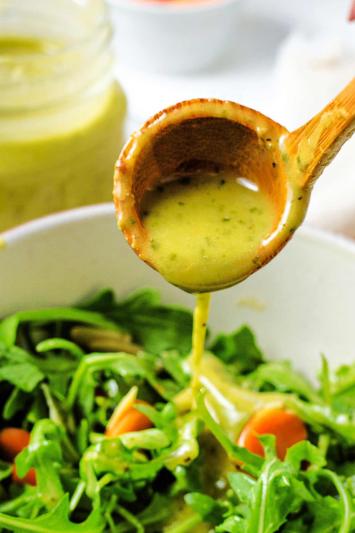 Drizzling lemon basil dressing out of a wooden ladle onto a bowl of greens on a table.