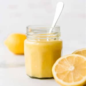 A jar of lemon curd with a spoon and sliced lemons on a table.